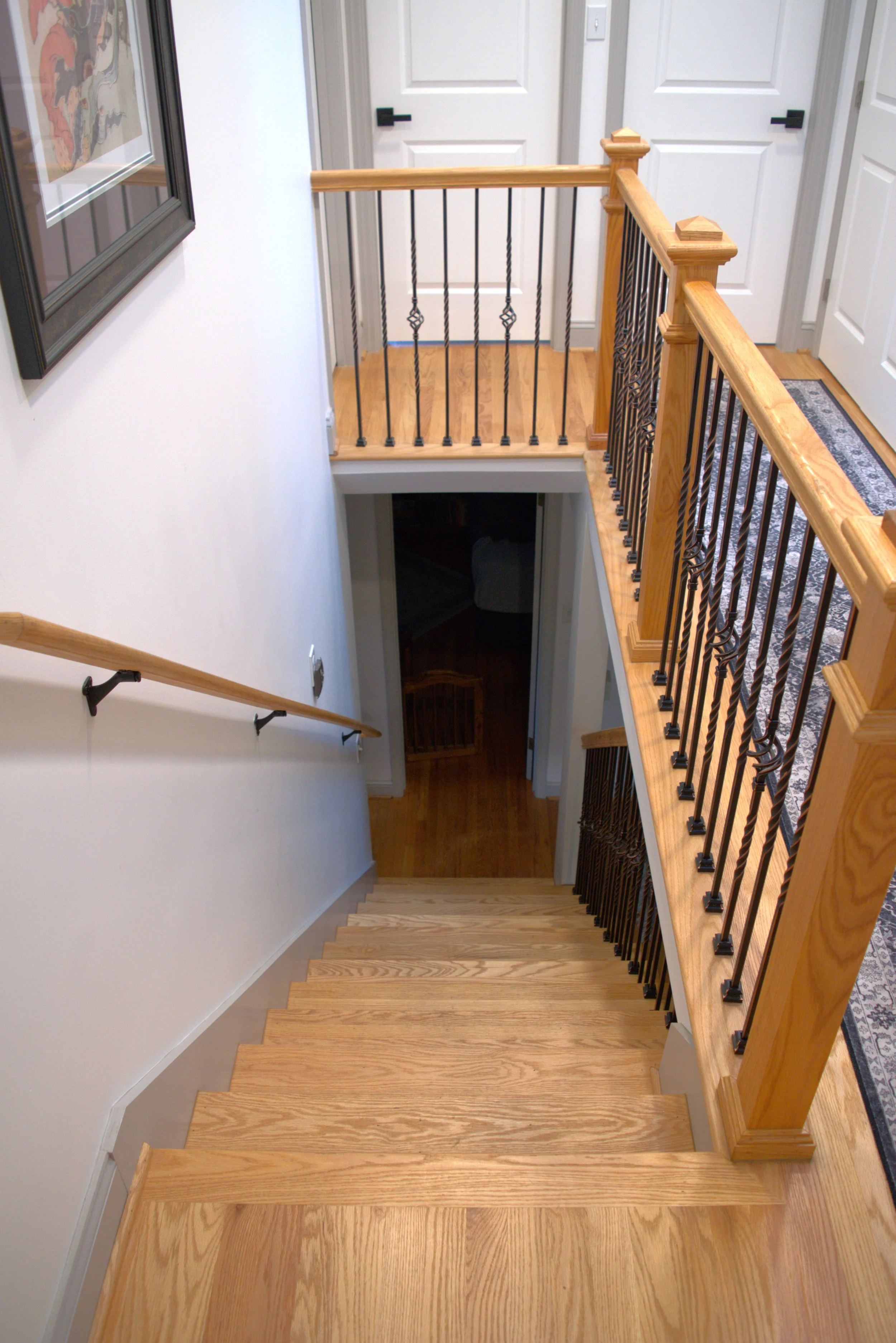 A view down a staircase with wooden steps and a decorative wooden and metal railing, leading to a downstairs hallway with a dark floor. The staircase is parallel to a white wall with a handrail, and there is a framed artwork on the wall. At the top o