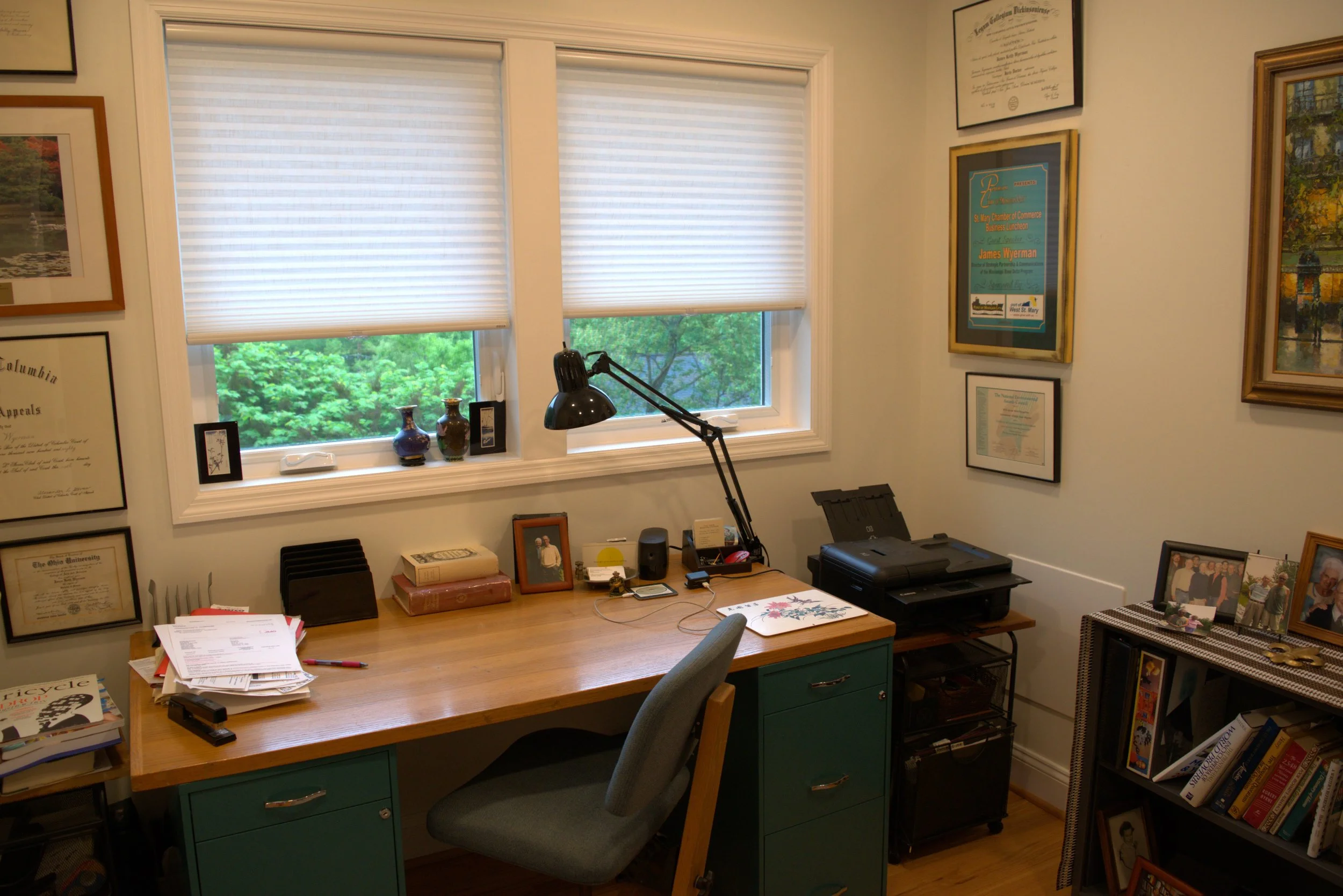 Office with a wooden desk, a gray office chair, shelves with books, framed certificates, and colorful decorations, and a window with blinds showing green trees outside.