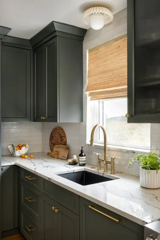 Modern kitchen with dark green cabinets, a white marble countertop, a black sink, and a window with wooden blinds. There are decorative items including a bowl of oranges, a wooden cutting board, a soap dispenser, and a potted plant.