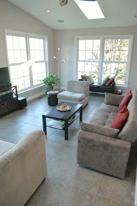 Bright living room with large windows, a beige sofa with red pillows, a black coffee table, a cream armchair, a small black side table, a TV stand with a TV, and a window seat with multiple pillows, sunlit with a view of tree branches outside.