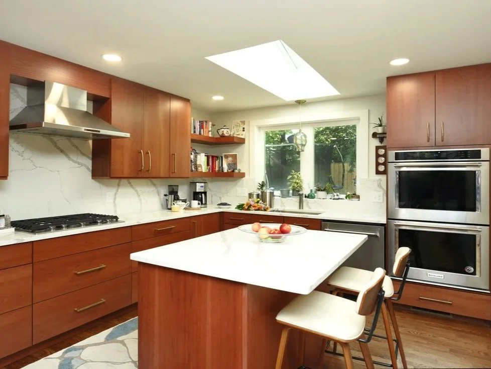 Modern kitchen with wooden cabinets, white marble countertops, a kitchen island with fruit, large window, built-in oven and microwave, skylight, and open shelves with books and decor.
