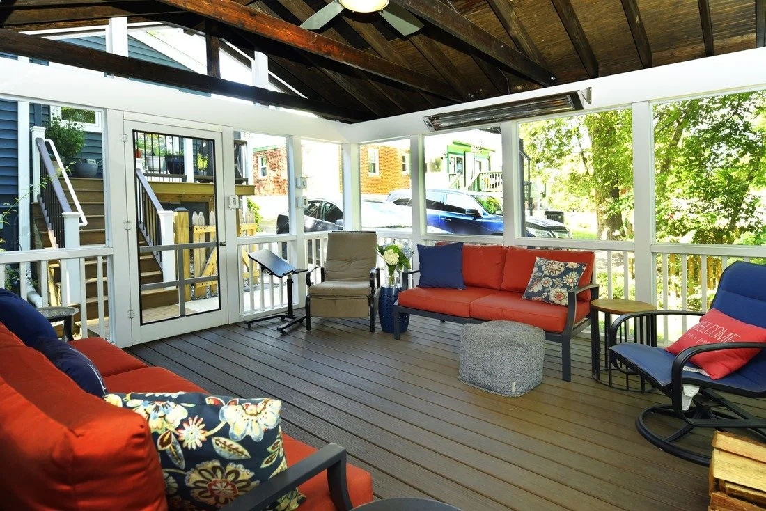 Sunroom with orange and blue cushioned chairs and sofa, wooden flooring, large windows, and outdoor view with trees and parked cars.