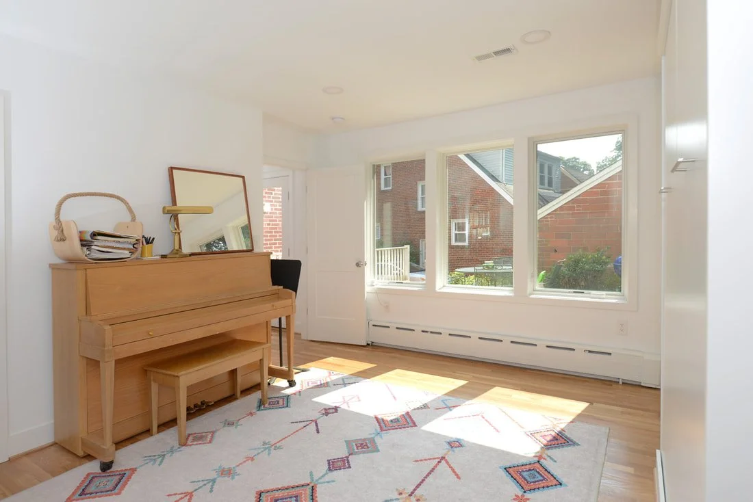 Bright living room with a wooden upright piano, a mirror, a chair, and a large window letting in sunlight.