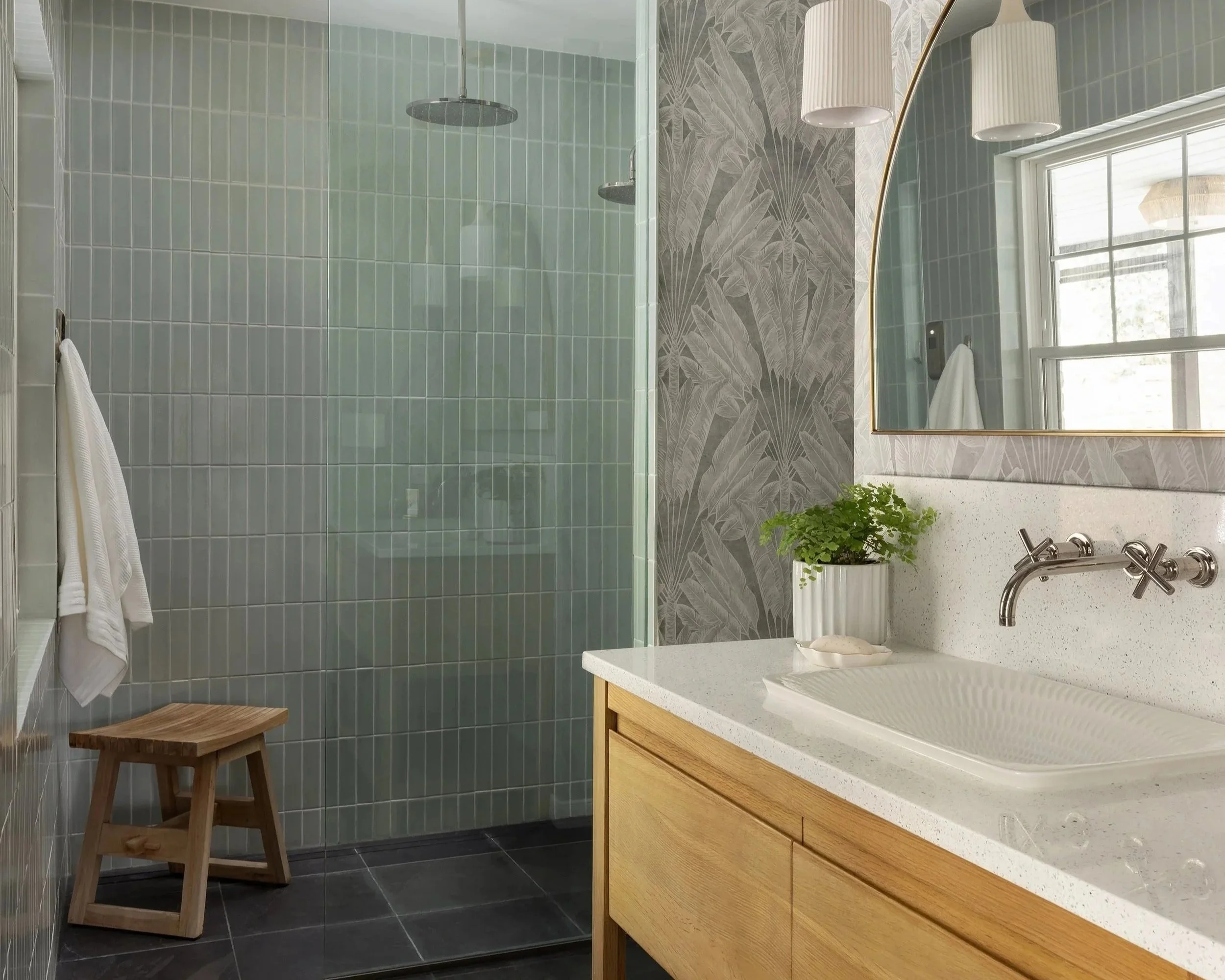 Modern bathroom with walk-in shower featuring green tiles, wooden stool, white towel, and vanity with a sink, mirror, and potted plant.