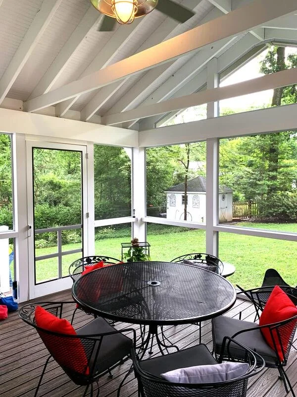 A screened porch with a black metal round table and five chairs with red and gray cushions. The porch has white wooden beams, glass windows, a door, and overlooks a green yard with trees and a small shed.