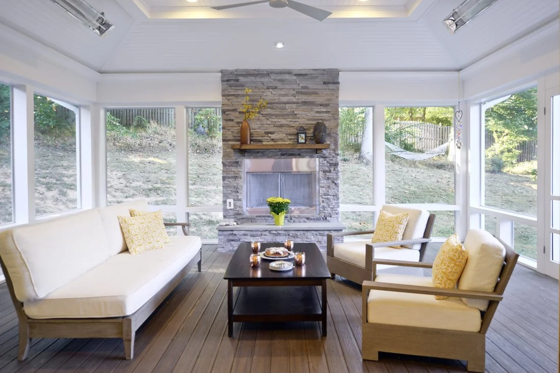 Sunroom with a stone fireplace, white ceiling, wooden floor, cream furniture, yellow pillows, and large windows with views of a backyard.