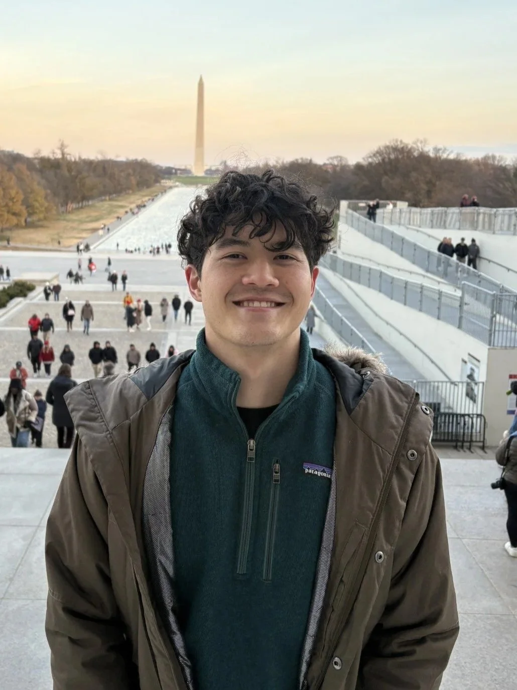 Smiling young man with curly hair in front of the Lincoln Memorial and the Washington Monument in Washington, D.C., during daytime.