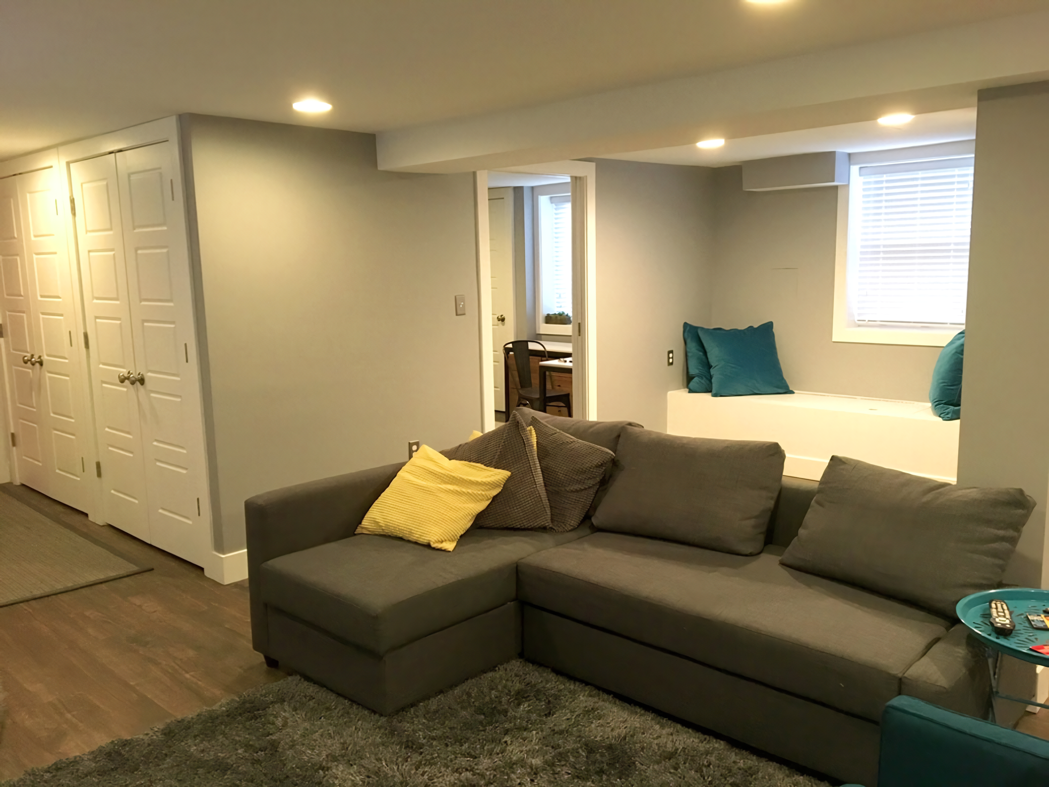 A living room with a gray sectional sofa with yellow and dark gray pillows, a window seat with teal pillows, a window with blinds, and a doorway leading to a small dining area with a table and chairs.
