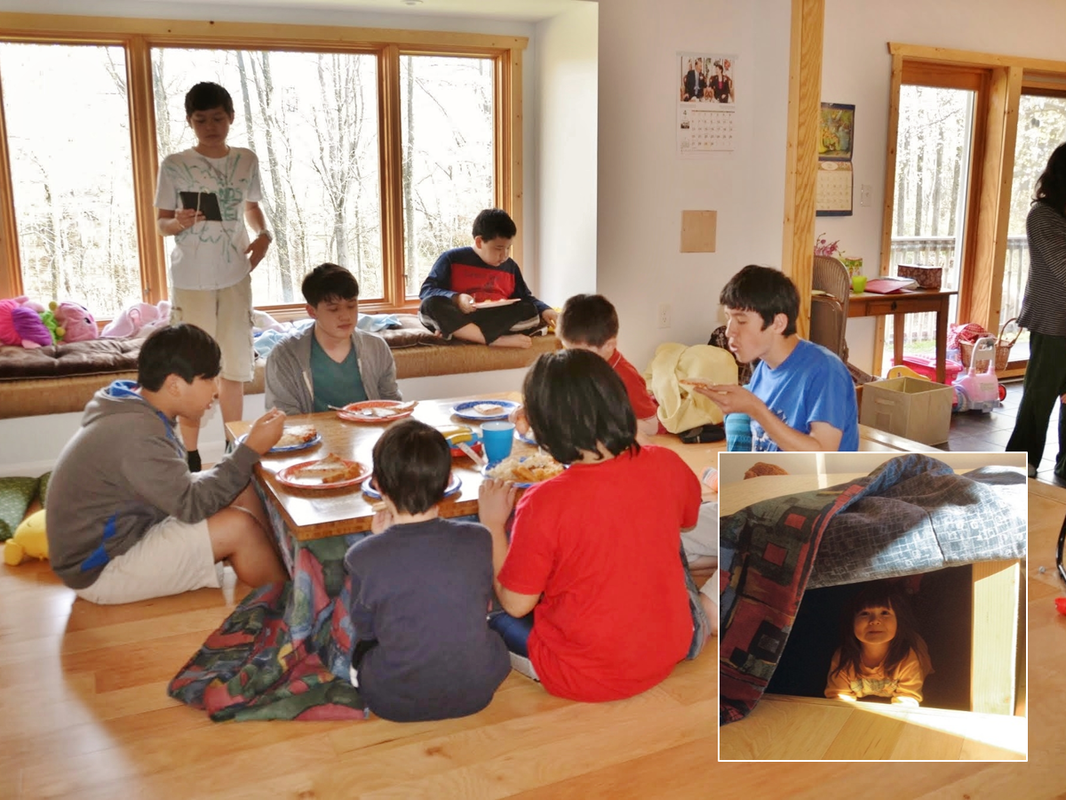 Group of children having a meal around a low table in a cozy room with large windows and natural lighting. Some kids are sitting on the floor, others are sitting on a bench, and one child is playing with a tablet in the background. An inset shows a girl inside a small makeshift tent under a blanket.