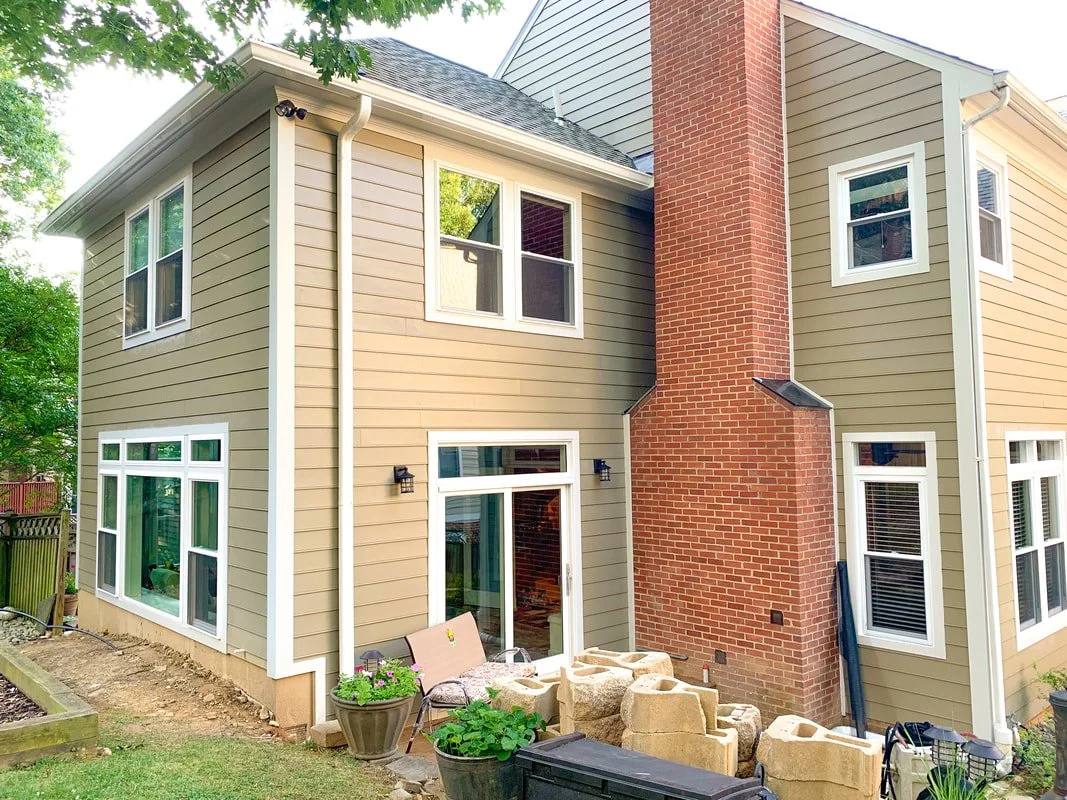 Rear view of a two-story house with beige siding, white trim, a brick chimney, multiple windows, and a sliding glass door leading to a backyard patio with garden plants, a bench, and concrete blocks.