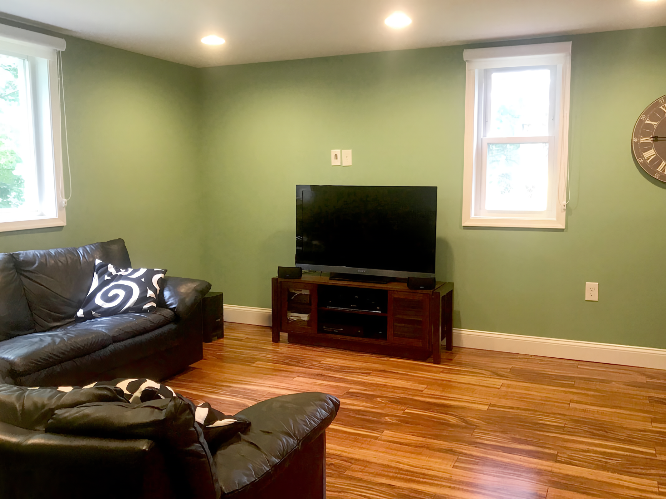 Living room with green walls, hardwood floors, two windows with white blinds, a black leather sofa with black and white pillows, a wooden TV stand with a flat-screen television, and a wall clock.