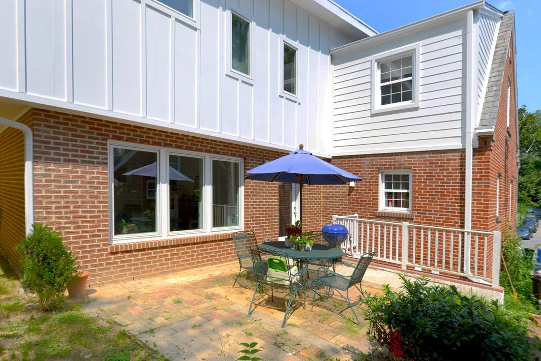 Backyard patio with brick and white siding on house, with outdoor table, chairs, umbrella, potted plants, and grill.