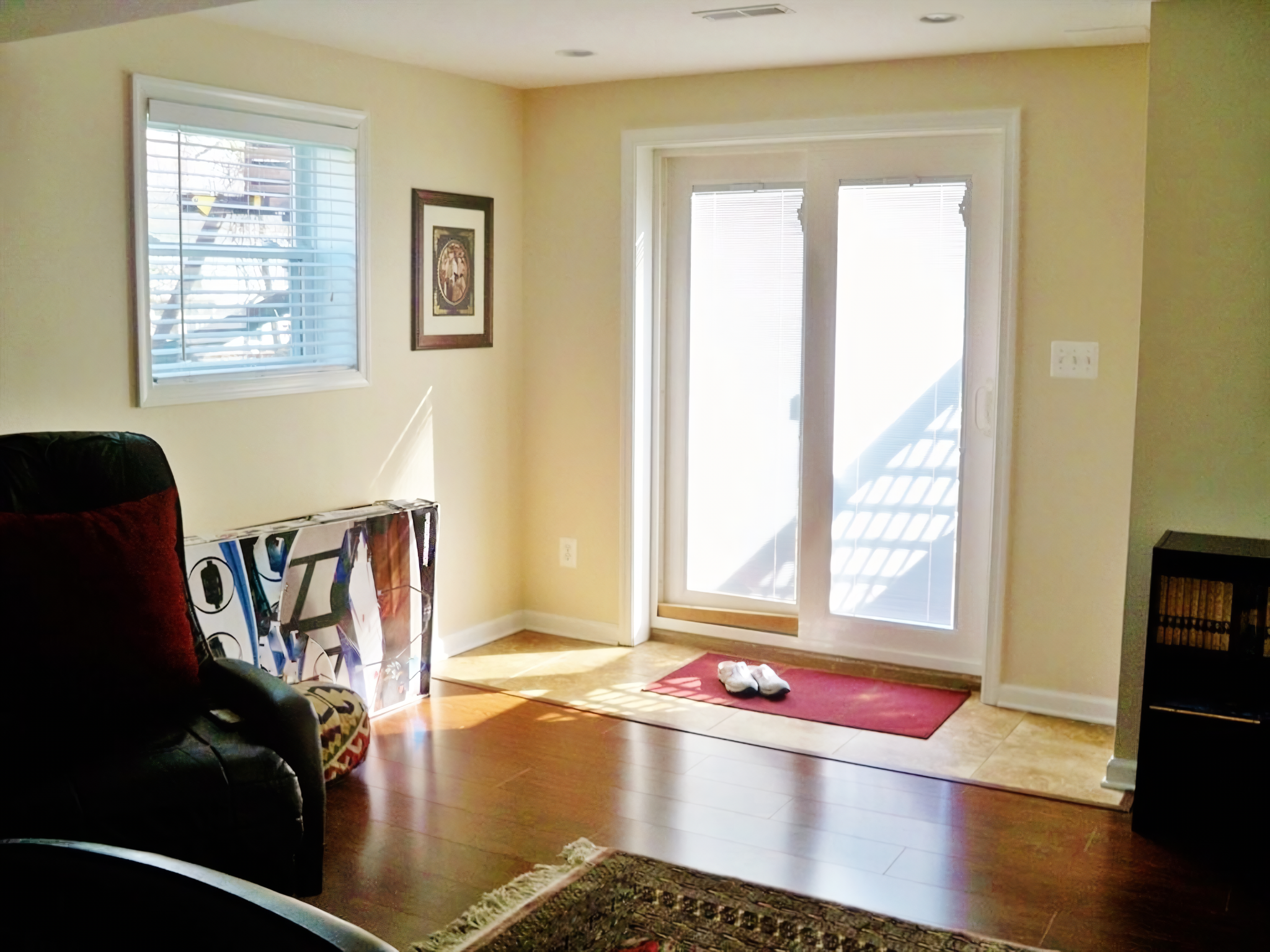 Living room with a window and sliding glass door leading outside, hardwood flooring, a framed picture on the wall, a red mat with a pair of white shoes, and a black chair with a red pillow.