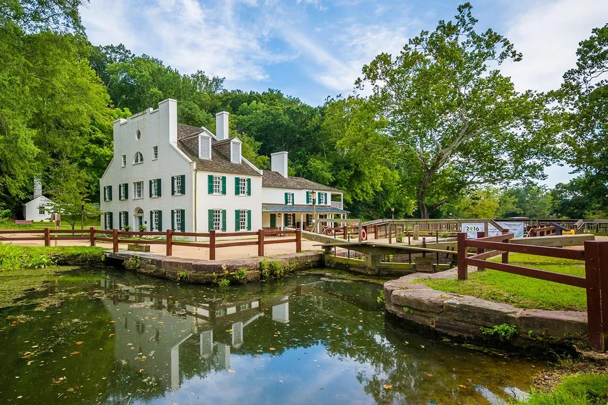 A large, historic white house with green shutters beside a pond, surrounded by greenery and trees.
