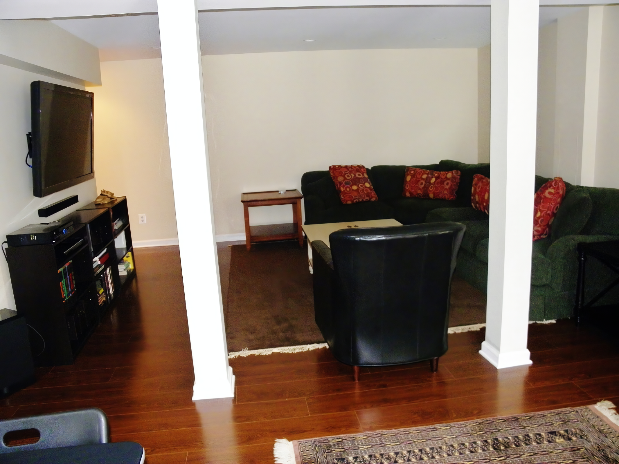 Living room with a large L-shaped green sofa with red patterned pillows, a black leather armchair, a wooden coffee table, and a flat-screen TV on a black entertainment center. The room has hardwood floors and a partial wall dividing the space.