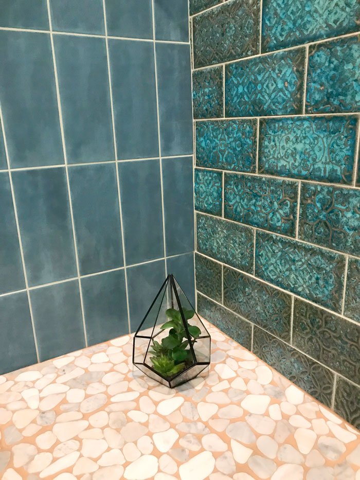 A small glass terrarium with green plants on a pebbled pink and white tiled surface, surrounded by blue tiled walls.