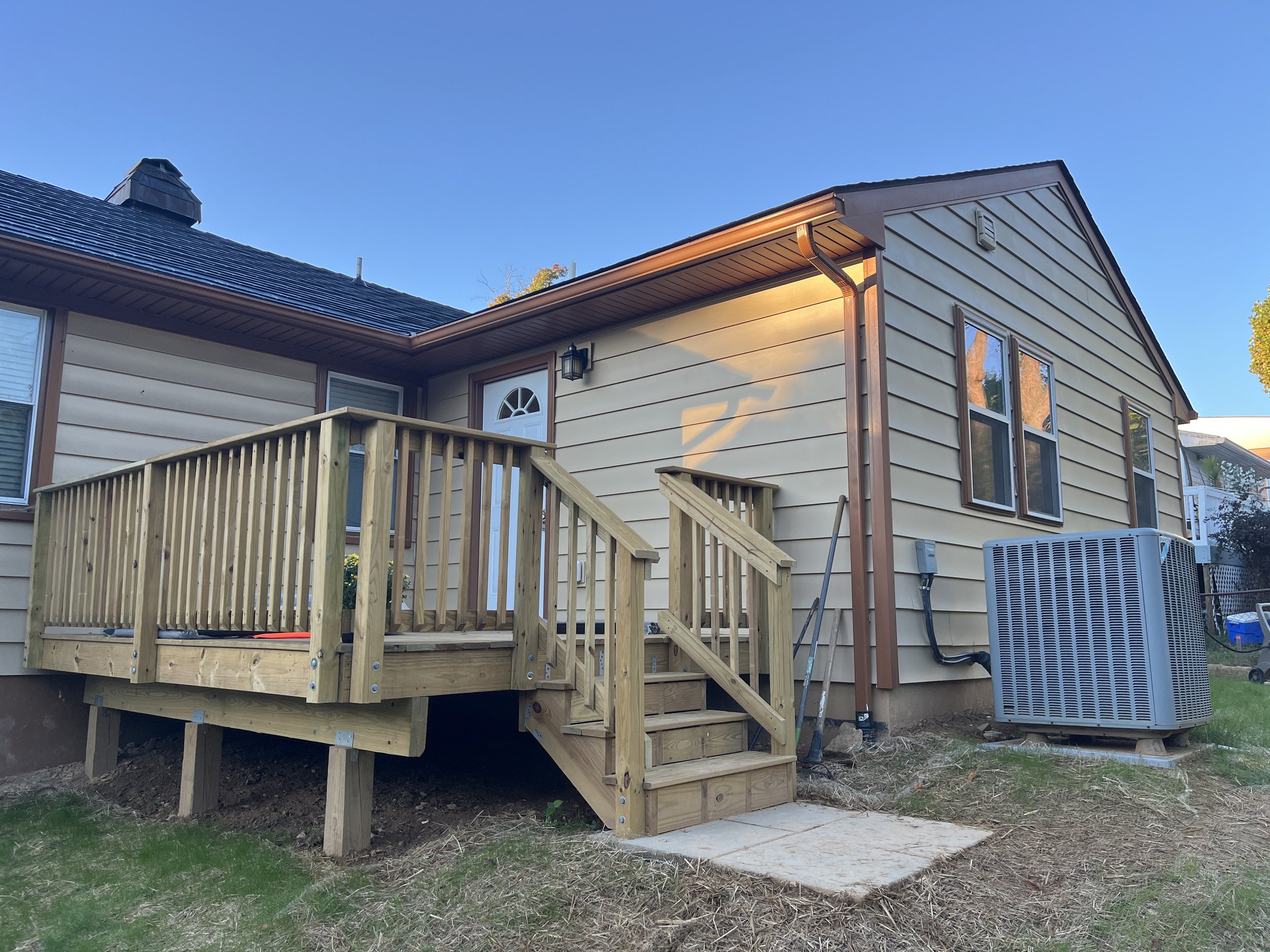 Backyard view of a two-story house with beige siding, a wooden deck with stairs, and a sliding glass door. There is an air conditioning unit and a large gray outdoor cooler outside. The sky is clear and blue.