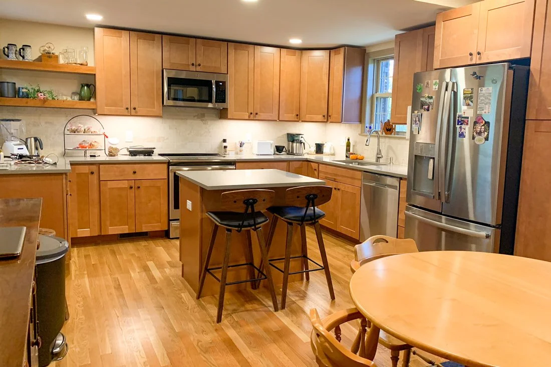 Modern kitchen with wooden cabinets, stainless steel appliances, a small kitchen island with two black bar stools, a round dining table with wooden chairs, hardwood floors, and a window above the sink.