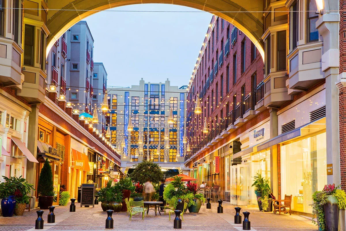 Outdoor shopping street with string lights, patio seating, potted plants, and storefronts at dusk.