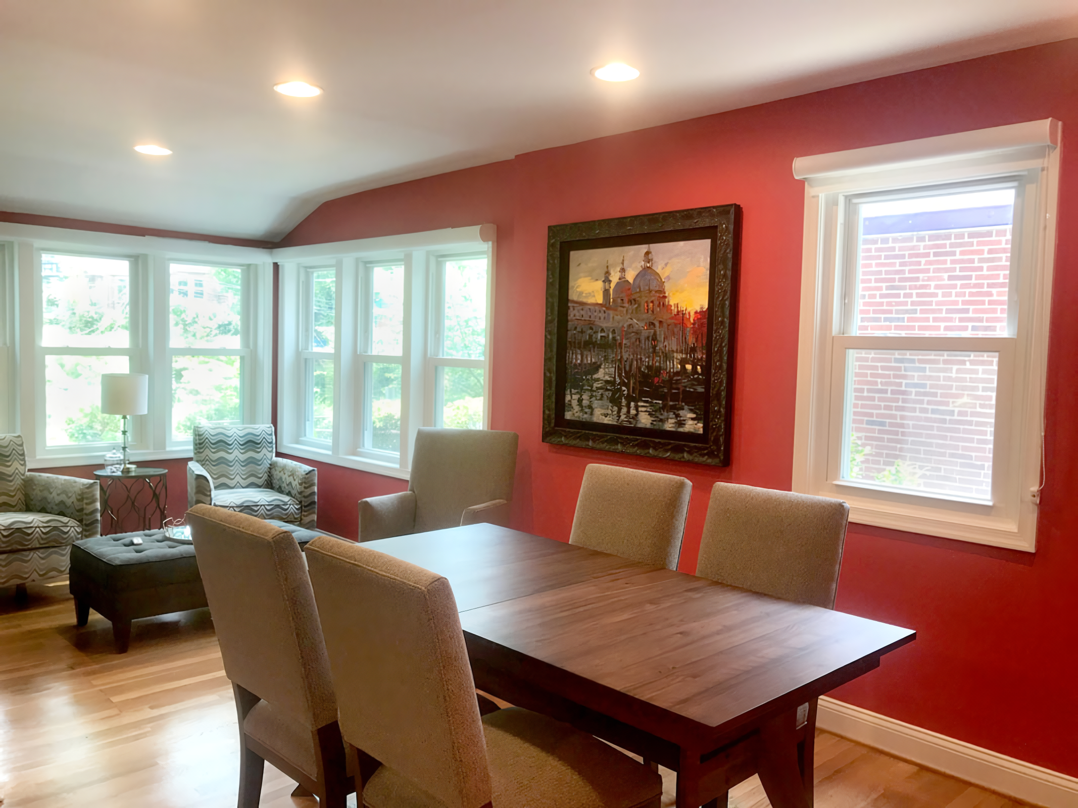 A dining room with a wooden table and beige upholstered chairs, a red accent wall with a framed painting, and multiple windows letting in natural light.