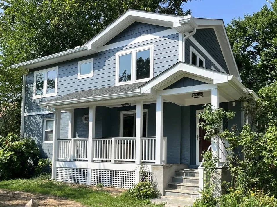 A two-story house painted in blue with white trim, featuring a front porch with white railing, steps leading up to the front door, multiple windows, and surrounded by green trees and bushes.