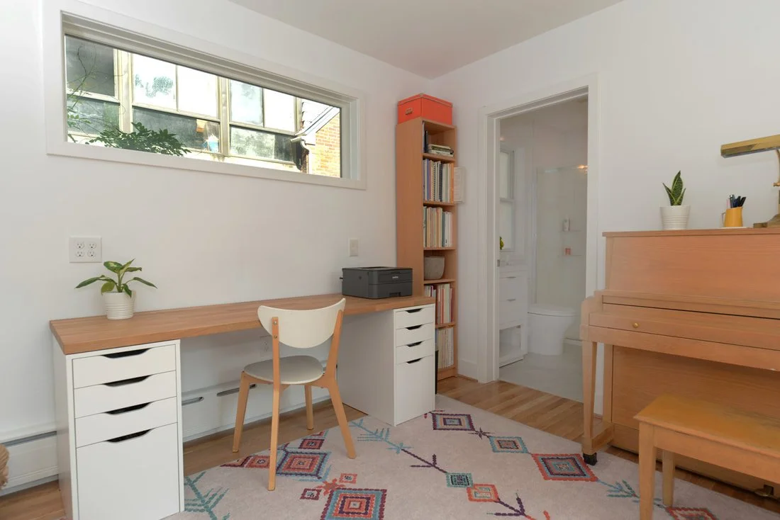 Home office with white walls, wooden desk with white drawers, white chair, potted plant, tall bookshelf with books and storage, wooden upright piano, colorful patterned rug, and a view of a bathroom through open door.