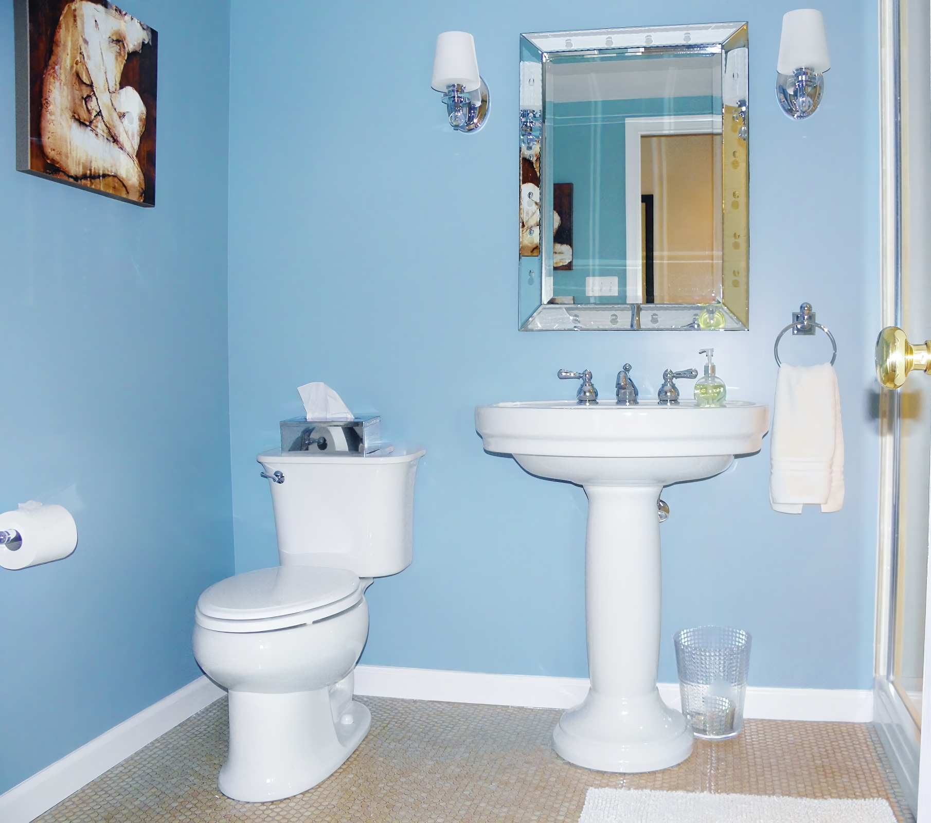 Bathroom with light blue walls, white toilet with tissue box on top, white pedestal sink with soap dispenser, mirrored wall cabinet, wall-mounted lights, beige tiled floor, white baseboard, and a painting on the wall.