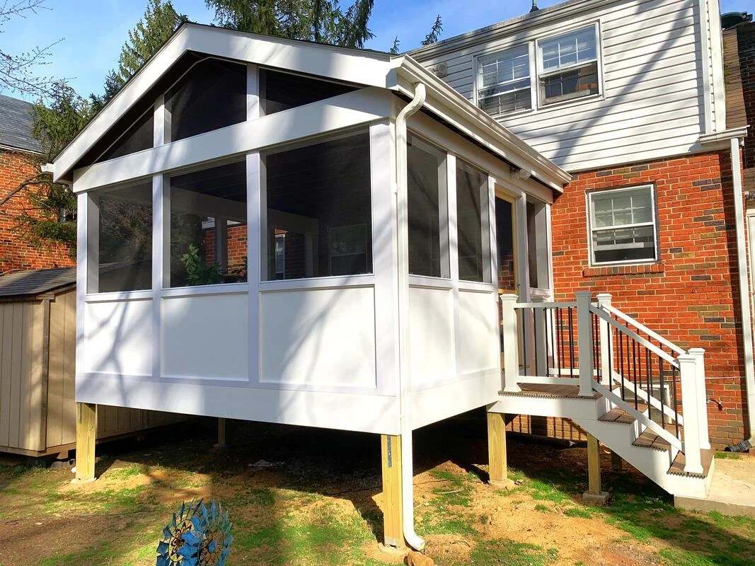 A newly built white screened porch with stairs attached to a brick house, surrounded by a backyard with grass and trees.