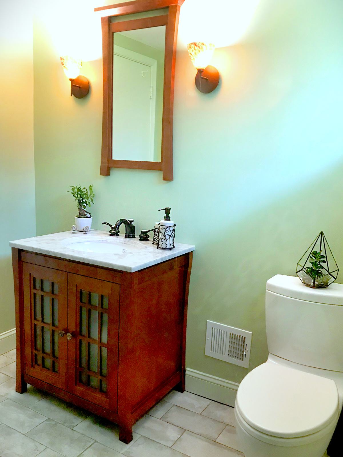 Bathroom with wooden vanity and marble top, mirror, wall sconces, potted plants, and a toilet with a geometric terrarium on top.