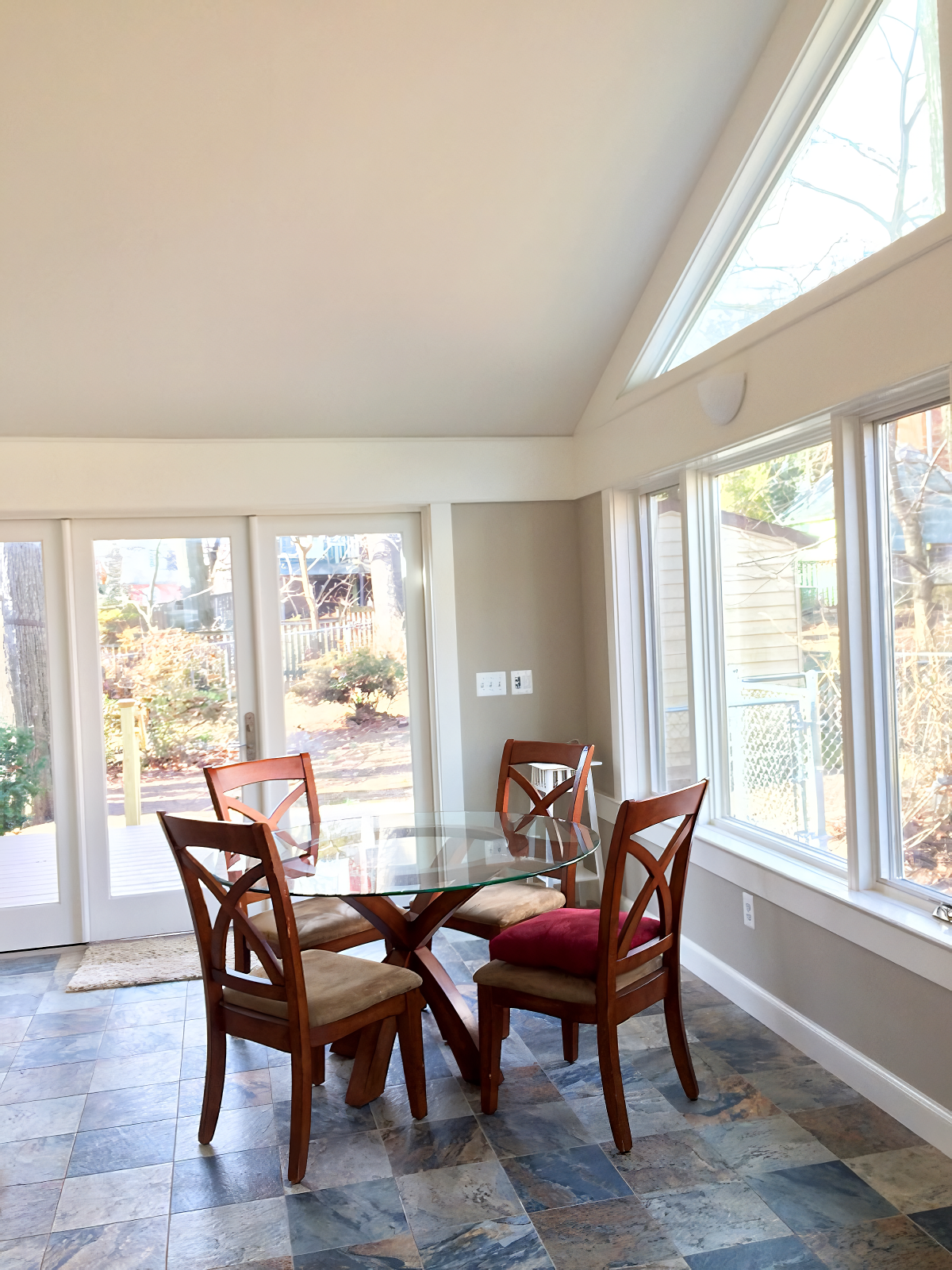Dining room with a glass-top table and four wooden chairs with beige and red cushions, surrounded by large windows and a sliding glass door leading to an outdoor yard, with a slate tile floor.