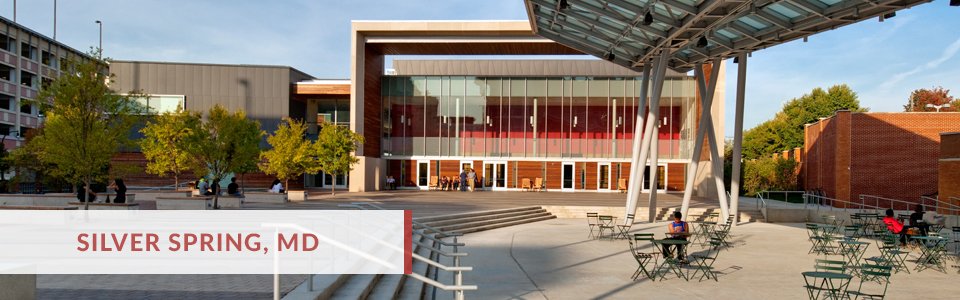 Exterior view of a modern building in Silver Spring, Maryland, with an open plaza featuring outdoor seating and a glass facade, trees, and a clear sky.