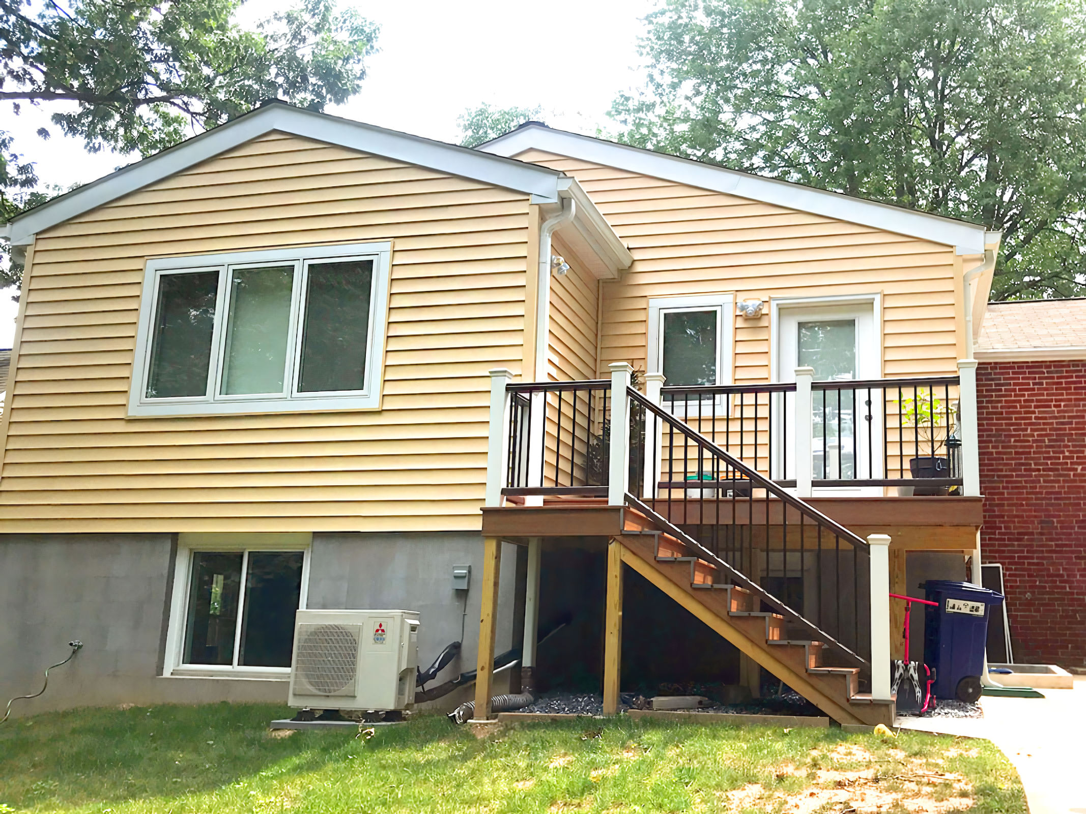 Back view of a two-story house with beige vinyl siding, white trim, and a small wooden deck with black railing. There are stairs leading to the deck, and a sliding glass door. Windows are on both floors. The house has an air conditioning unit outside