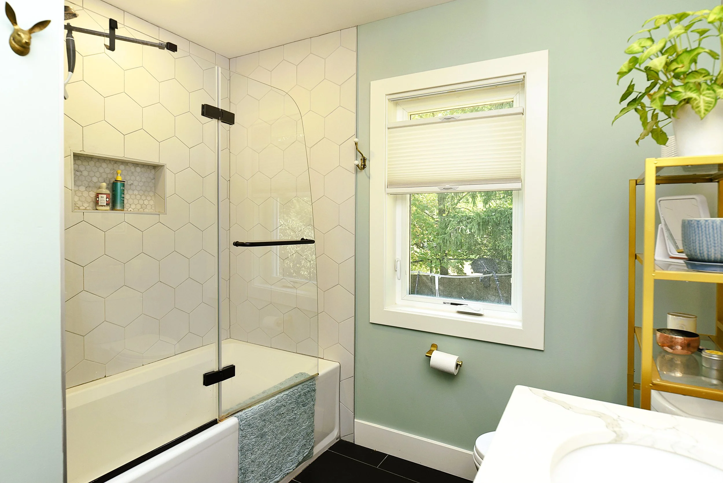 Bathroom with a walk-in shower enclosed with glass, hexagon tile on the wall, a window with a view of greenery, a yellow shelving unit with toiletries, a towel hanging on the shower, and a toilet paper roll.