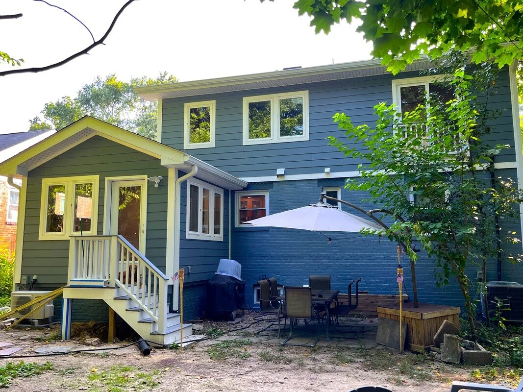 Backyard of a house with blue siding, white trim, a small staircase, outdoor table with umbrella, and trees.