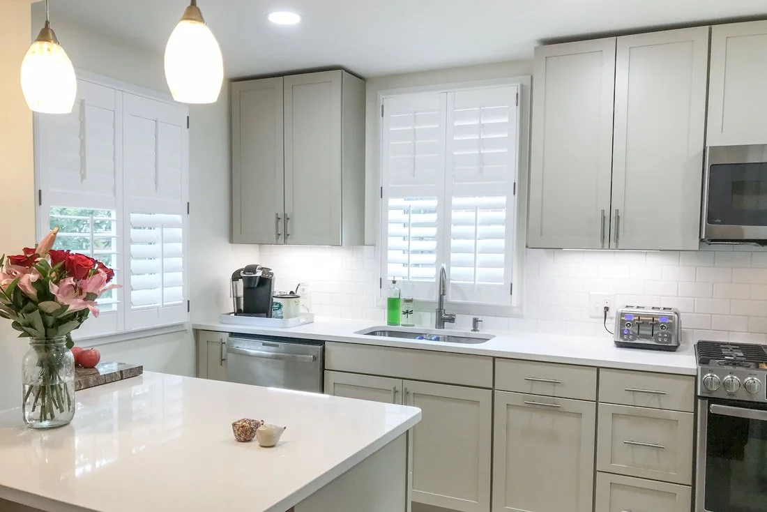 Modern kitchen with white cabinets, white countertops, stainless steel appliances, a window with shutters, a vase of pink and red flowers on the island, and pendant lights.