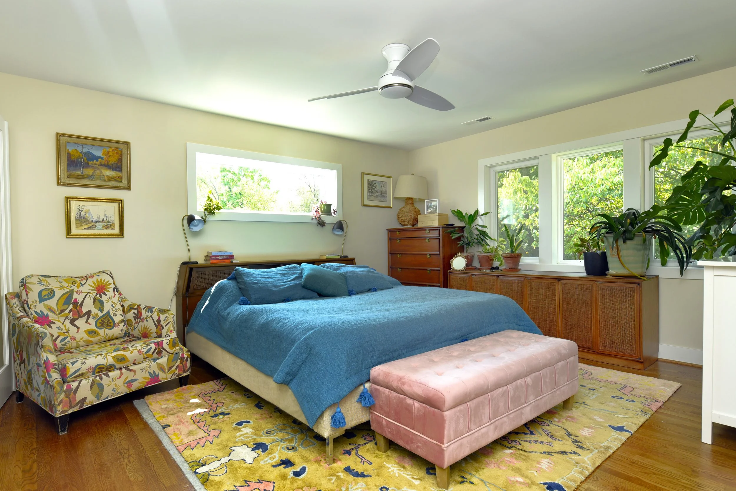 A cozy bedroom with a blue bedspread, a pink velvet bench at the foot of the bed, and a vintage floral armchair. There are large windows with greenery outside, wooden floors, and floral and landscape artwork on the walls. Potted plants are on a woode