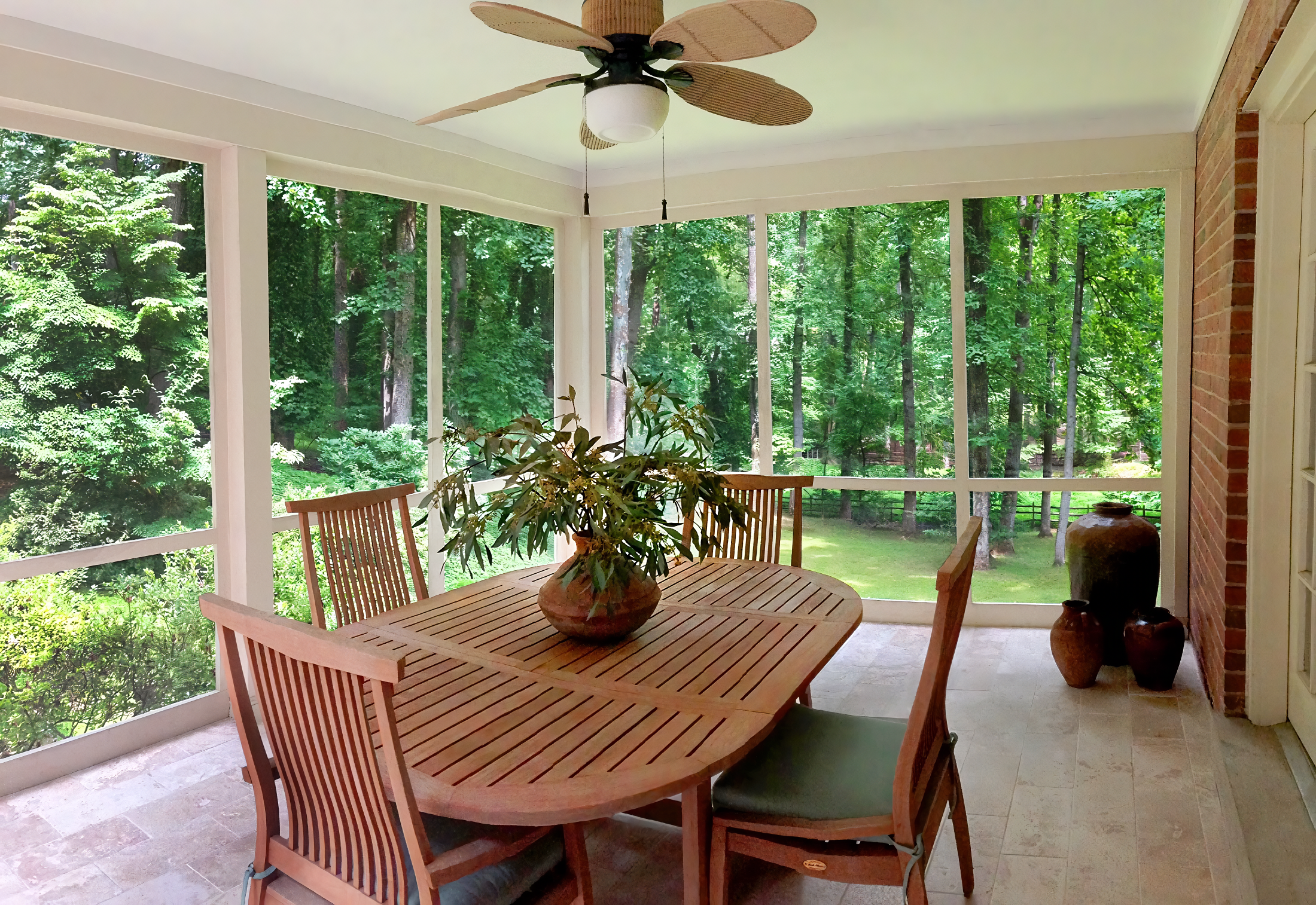A screened porch with a wooden dining table and chairs, a ceiling fan, and decorated with large pottery vases, overlooking a lush green forest.