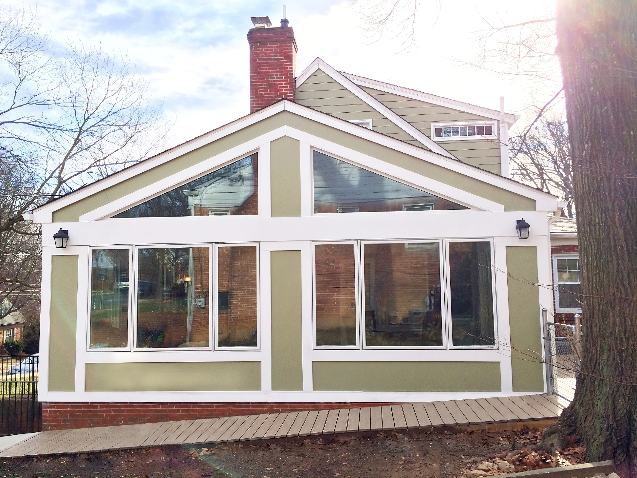 A house with a green structure attached to the back, featuring large windows and a gable roof with white trim, surrounded by trees and a wooden deck in the yard.