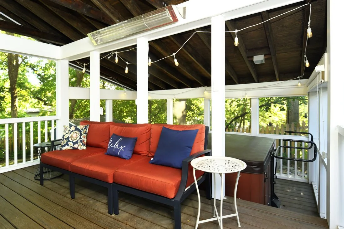 Screened porch with orange cushion sofa, blue pillows, small white side table, hot tub, string lights, and a heater on the ceiling, surrounded by trees.
