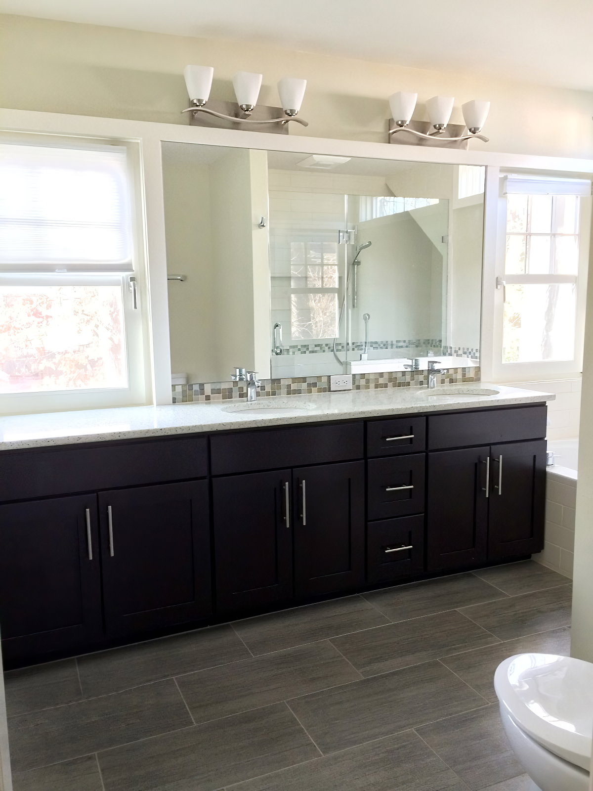 Bathroom with double sink vanity, dark wood cabinets, granite countertop, large mirror, modern light fixture, window, and a walk-in shower visible in the reflection.
