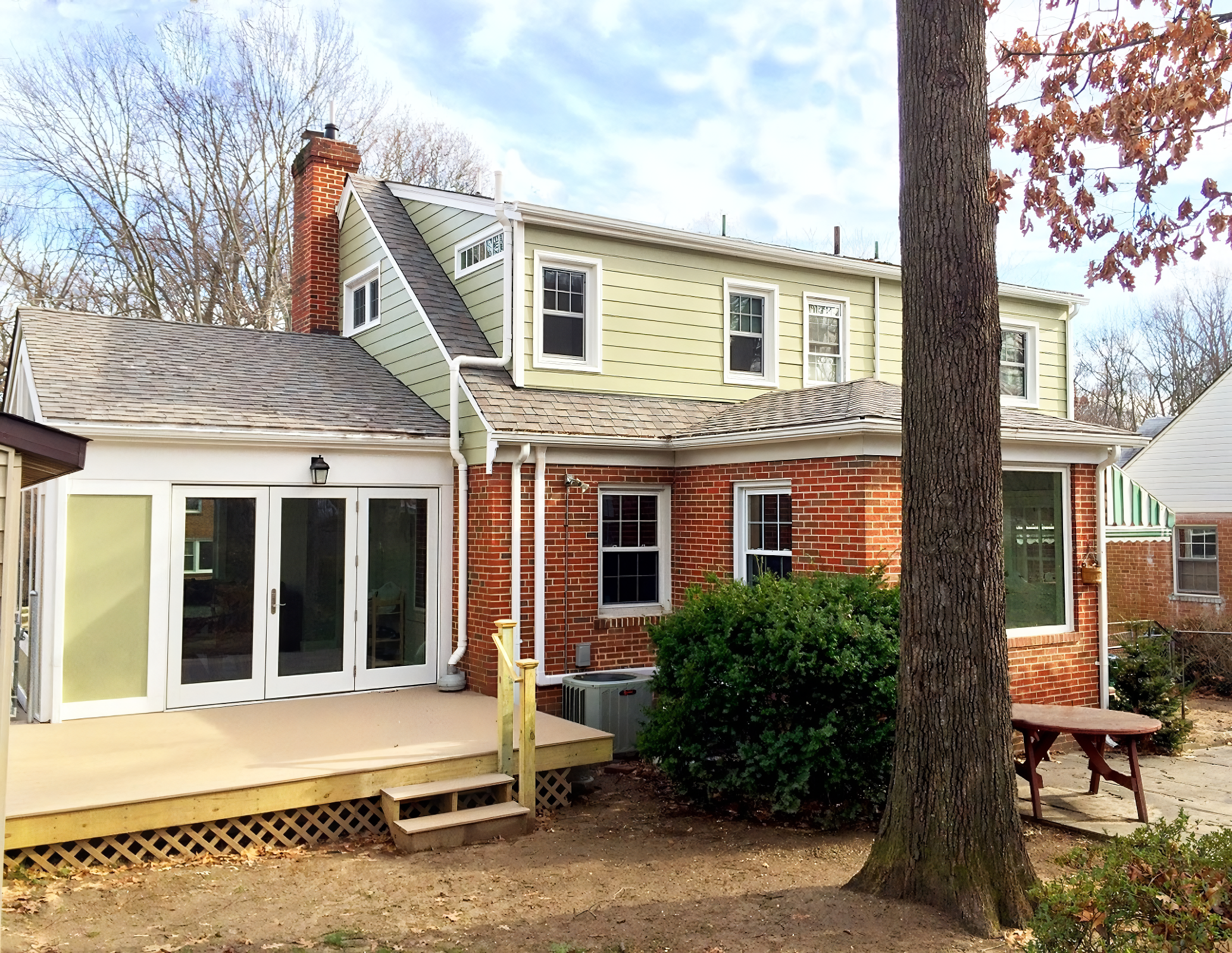 Back view of a house with a small wooden deck, bushes, a large tree, and an outdoor picnic table, with a partly cloudy sky in the background.