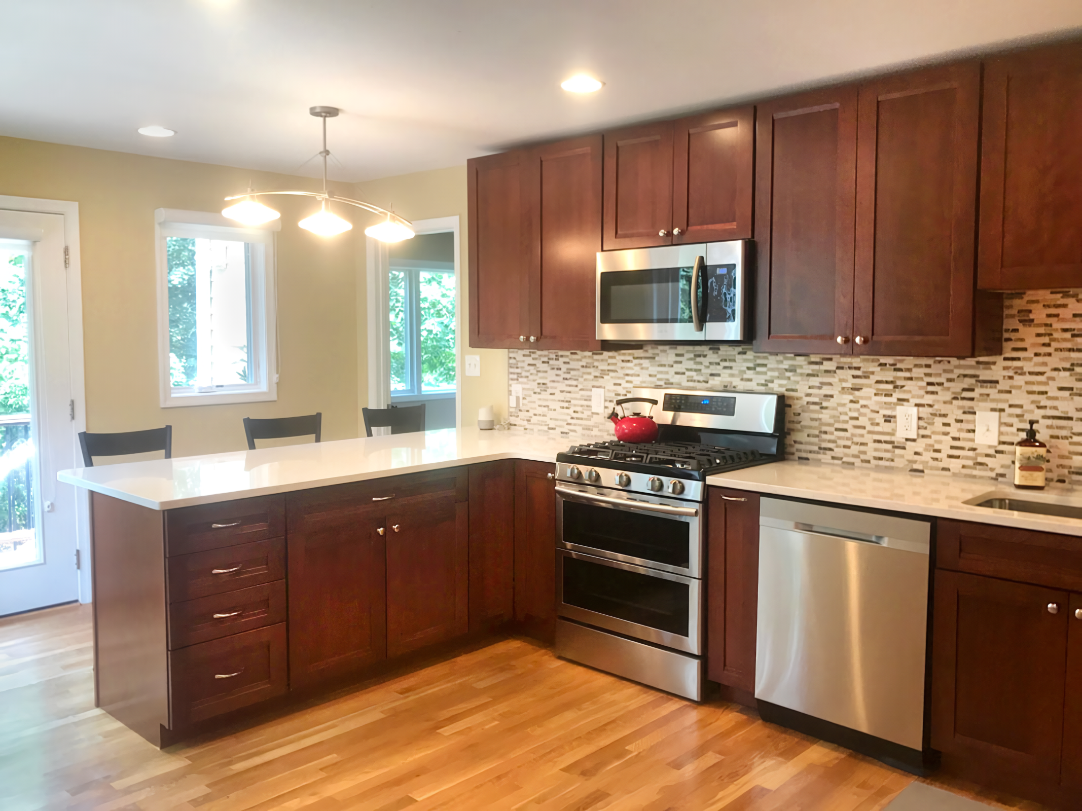 A modern kitchen with wooden cabinets, a stainless steel stove and microwave, a mosaic tile backsplash, and a white countertop island with three black chairs, illuminated by ceiling lights and natural light from windows and a glass door.