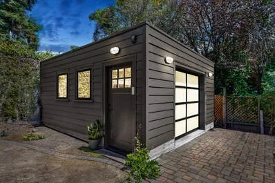 A small dark gray modern garage with a white door and windows, located in a backyard with trees and a paved driveway.