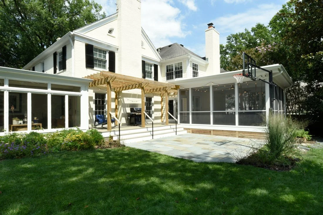 Backyard view of a large white house with a screened porch and a wooden pergola, featuring a basketball hoop, a grill, and a landscaped lawn with trees and bushes.