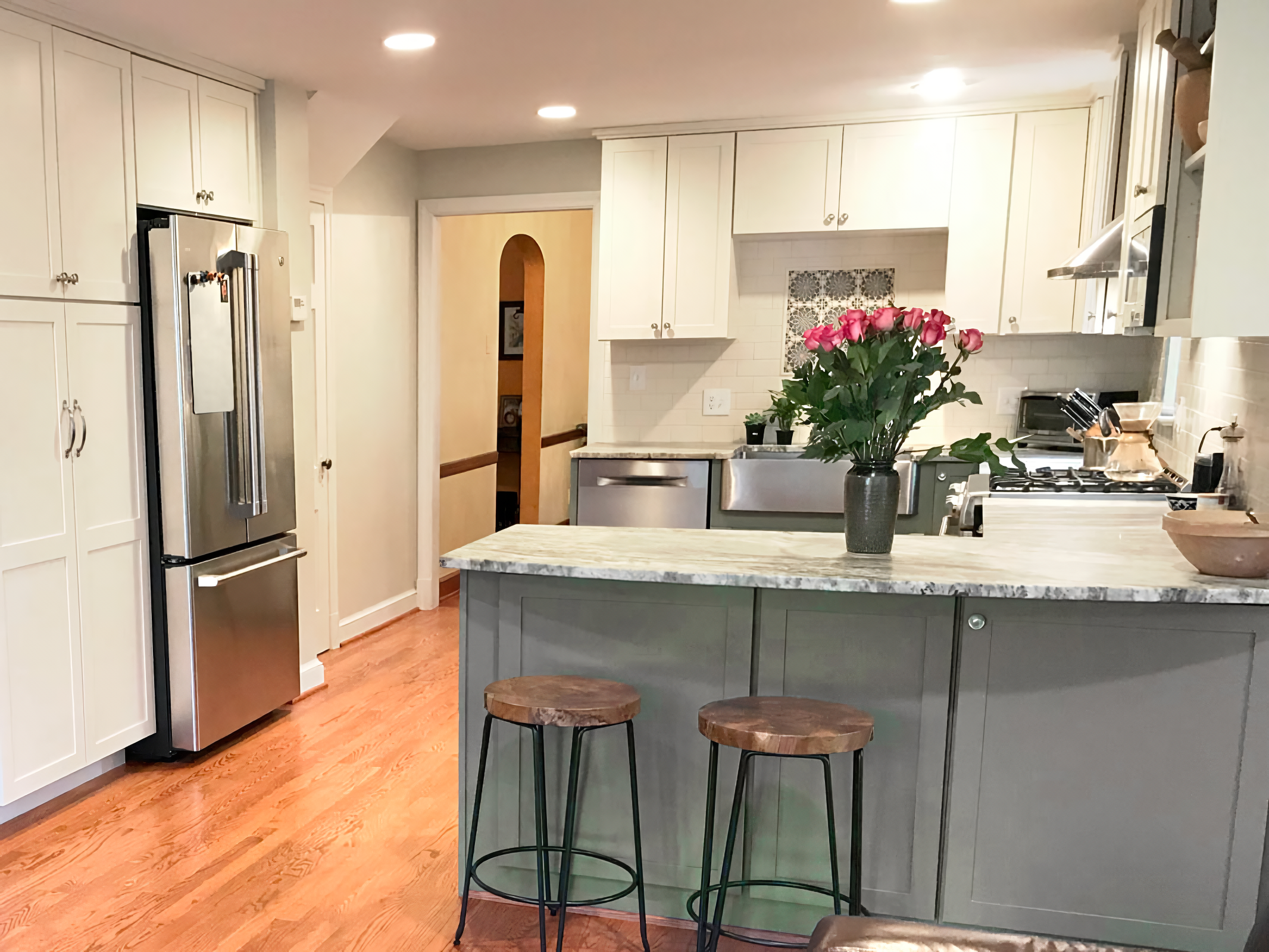 A modern kitchen with white cabinets, stainless steel appliances, a marble countertop island with two wooden stools, and a vase of pink roses on the counter.