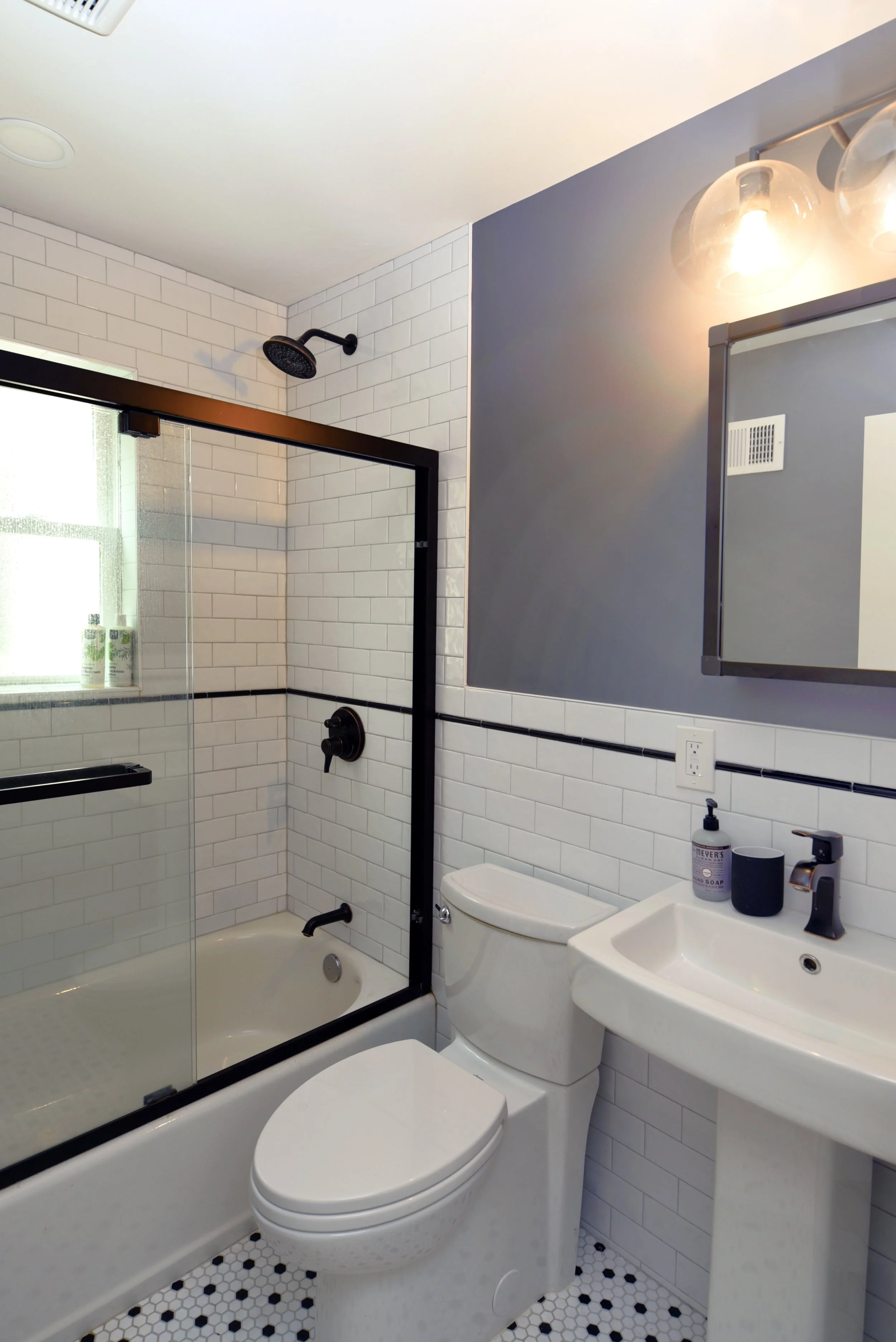 Small modern bathroom with white subway tile walls, a glass shower door, a white toilet, a small sink, and a mirror with light fixtures above.