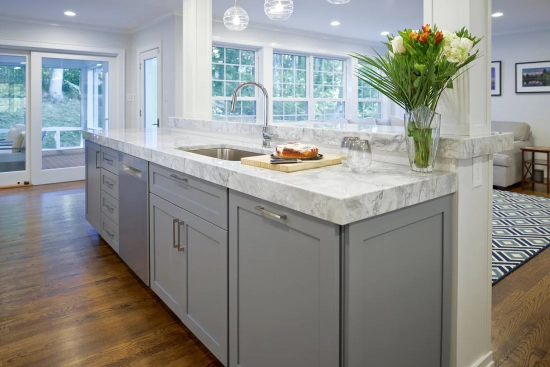 Modern kitchen with white marble countertops, gray cabinets, a vase of flowers, and a view of a living room with large windows and a sliding glass door leading to a balcony.
