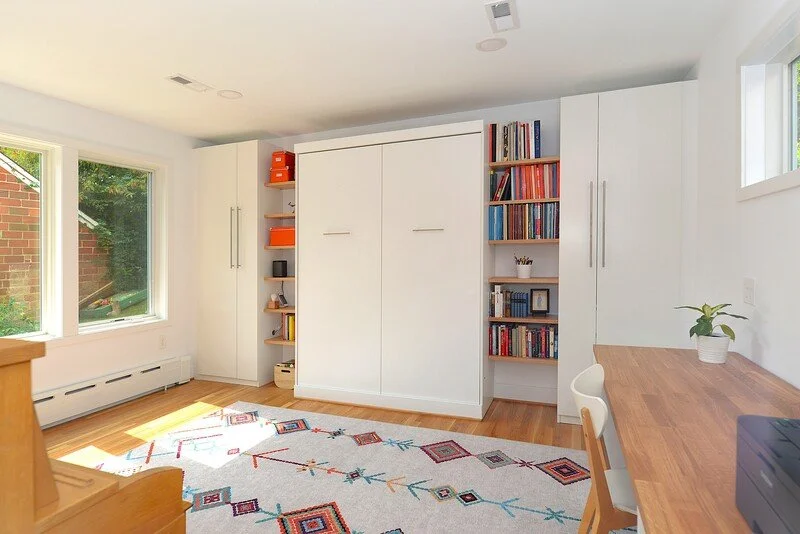 Bright home office with white built-in shelves and cabinets, a wooden desk with a white chair, a potted plant, a colorful geometric rug, large windows, and hardwood floors.