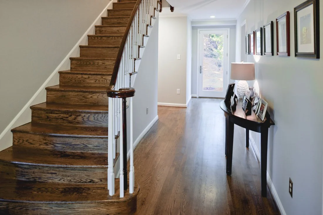 Empty hallway and staircase in a home with light-colored walls, wooden flooring, a small table with framed photos and a lamp, and a door leading outside.