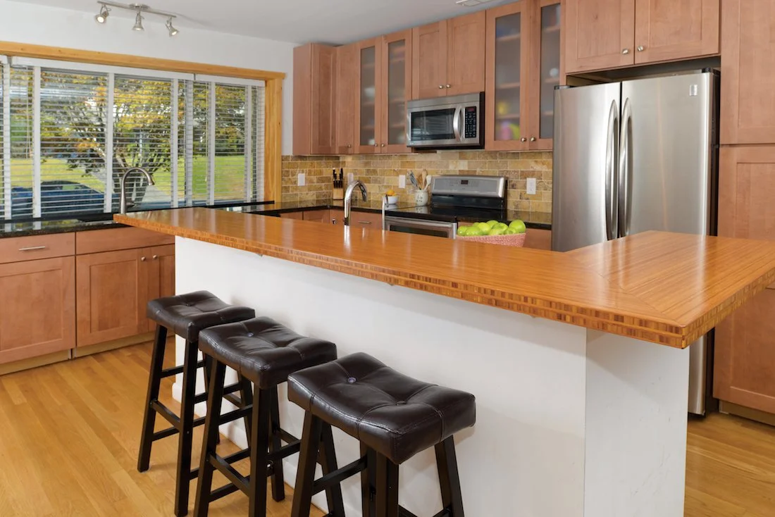 Kitchen with wooden cabinets, a stainless steel refrigerator, microwave, and an island with three black bar stools.